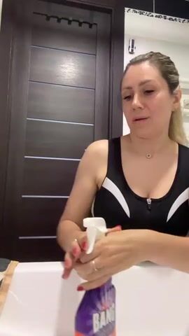 A woman with blonde hair pulled back in a ponytail is in a bathroom, wearing a black and white sports bra. She is holding a spray bottle and appears to be cleaning a white sink. The bathroom has a dark wood door and a mirror. The woman is speaking.