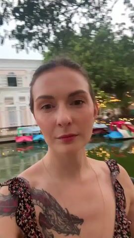A woman with a dragon tattoo on her chest and arm smiles at the camera. She is standing outdoors at dusk near a body of water with swan boats and string lights. She is wearing a floral dress.