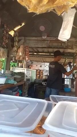A person wearing a black t-shirt and a baseball cap is seen preparing food in what appears to be an outdoor food stall. The stall has a corrugated metal roof and is filled with various cooking equipment and supplies, including a fan, shelves stocked with items, a stove, and large food containers in the foreground. The person is actively engaged in cooking or serving, using utensils and interacting with pots and containers. The overall environment suggests a busy, functional food service operation.