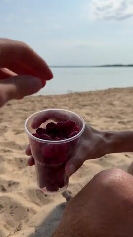 A close-up shot shows a person's hands reaching into a clear plastic cup filled with raspberries. The cup is held in the person's left hand while their right hand plucks a raspberry. The background consists of a sandy beach and a body of water under a bright sky, suggesting an outdoor setting. The focus is on the interaction with the raspberries and the hands.