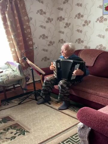 An elderly white male with short white hair and wearing a dark blue jacket and camouflage pants sits on a dark red couch and plays an accordion. He is holding sheet music on a stand in front of him and there is a microphone attached to the stand. The room has floral wallpaper and a rug on the floor. There are two other couches in the room, one in the foreground and one partially visible to the right. A window with brown curtains is to the left of the man.
