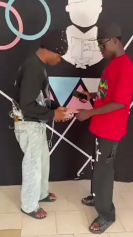 Two young men stand facing each other in front of a decorated wall. One is wearing a black beanie, a black long-sleeved shirt, light blue patterned jeans, and black sandals. He also has a cross-body bag. The other man wears a black baseball cap, glasses, a red t-shirt, black pants with white cross accents, and black sandals. They appear to be in a disagreement. The man in red places his hand on the other's shoulder, and they then gesture and talk emphatically with their hands. Both hold smartphones during the interaction. The wall behind them has a black background and is decorated with colored circles, triangles, white lines, and a white cartoon drawing of a person reading a book labeled "Holy Bible." The floor appears to be light-colored tile.
