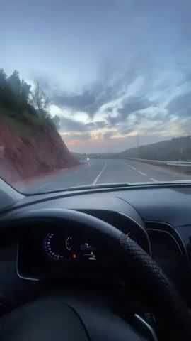 The view from inside a moving car driving on a winding road at dusk or dawn is depicted in the video. A red soil embankment sits to the left of the road. A few motorcycles and one or two cars are parked on the shoulder of the road at the curve. The sky is covered with scattered clouds, and a hint of orange is visible on the horizon. The interior of the car features a black steering wheel with a textured cover and a dashboard displaying the car's information. The overall atmosphere is calm and scenic.