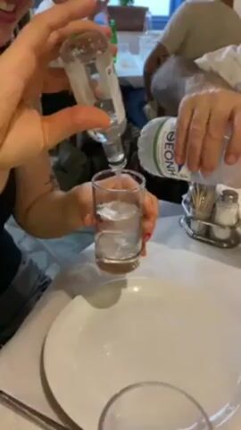 Here is a detailed description of the video:

The video captures a person pouring a clear liquid from a small glass bottle into a shot glass already partially filled with ice cubes and liquid. Concurrently, another person pours water from a plastic bottle labeled "DEONHA" into the same shot glass. This mixing of liquids occurs at a white-tablecloth-covered table in what appears to be a restaurant or dining area.  

The shot glass is adorned with a "15CL" marking.  Around the table, additional people can be seen dining. A small salt and pepper shaker set is also present on the table. The overall setting indicates a casual dining experience, possibly during a meal.