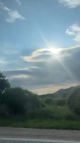 The video shows a slightly shaky, horizontal shot of a roadside with grass and trees against a bright sky with clouds. The sun is visible through some of the clouds, casting rays of light. The camera appears to be moving slightly, possibly from a vehicle. No people or specific objects are clearly visible beyond the natural environment. The time stamps indicate the video lasts about 4 seconds.