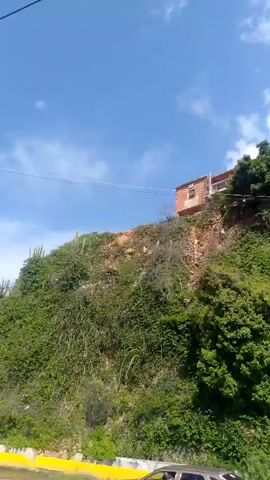 The video pans across a steep, overgrown hillside under a bright blue sky with scattered white clouds. Several houses, appearing to be made of brick and some with corrugated metal roofs, are situated precariously on the upper edge of the slope. The hillside itself is covered in dense green vegetation, including trees and vines. Some areas of exposed, reddish-brown soil are visible, suggesting erosion or unstable ground. A yellow line is partially visible at the bottom left corner, possibly indicating a road or pathway. A utility pole with electrical wires stands on the right side of the frame, further up the hill. The overall impression is one of a rural or semi-rural setting with homes built on a challenging and potentially unstable terrain.