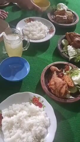 The video shows a top-down view of a meal spread out on a green surface, likely a tablecloth or mat. Several plates of white rice are visible, along with dishes containing fried chicken, cucumber slices, and leafy greens. There's a glass of iced yellow liquid with a straw, and a blue bowl. In the background, a person's hand is reaching for a small yellow bowl. Another person's feet are also visible at the edge of the frame. The lighting suggests it is nighttime or an indoor setting. The overall impression is of a casual shared meal.
