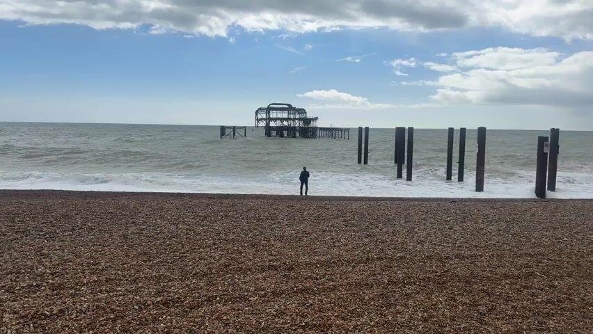 The scene is a beach with a person standing at the edge of the water, facing the sea. The beach is covered in pebbles. In the distance, there's a dilapidated pier structure in the water. To the right of the person, there are several wooden posts sticking out of the water. The sky is cloudy with patches of blue. The sea has moderate waves. The person is facing away from the camera.