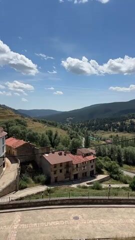 This is a vertical, handheld video shot from a slightly elevated position, looking out over a village nestled in a valley. The sky is a clear blue with scattered white cumulus clouds. In the foreground, there is a paved courtyard with a decorative brick pattern. A low wall with a metal railing separates the courtyard from a sloping grassy area. Beyond this, several buildings with red tile roofs and stone facades are visible, some appearing to be older village houses. A road winds through the middle ground, bordered by trees and fields. The background is dominated by rolling hills covered in dense green forest, with a few scattered buildings visible on the slopes. The overall impression is a peaceful, rural landscape on a sunny day.