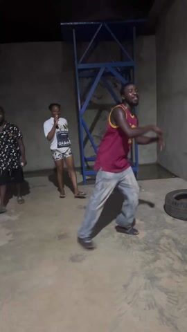 Here's a concise description of the video:

Two young adults are dancing indoors, with a third person partially visible on the left. The man is wearing a maroon and yellow basketball jersey and gray pants. The woman is wearing a white t-shirt and patterned shorts. They are smiling and seem to be enjoying themselves. 

The setting appears to be a concrete-floored room, possibly a garage or unfinished space, with gray walls. A blue metal structure stands in the background, along with a tire. The lighting is dim.