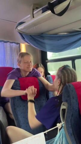 The video shows a young, blond-haired woman wearing a purple t-shirt, smiling and leaning over the back of a bus seat to interact with another young woman wearing a dark shirt and sunglasses, who has her hair in braids. A third young woman is visible behind the blond woman, seated in the bus seat. The interior of the bus includes blue-patterned seat upholstery, red headrest covers, blue curtains, and a white overhead compartment. The women appear to be having a friendly interaction, holding hands and smiling. The bus window is visible in the background. The song in the background is in Russian.