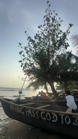 The video captures a serene beach scene, likely at sunrise or sunset due to the bright, warm sunlight reflecting off the water. Several wooden fishing boats are pulled up onto the sandy shore, with gentle waves lapping the coastline in the background.

In the foreground, a dark-colored wooden boat is prominently featured, with the white painted text "NOTHING PASS GOD" visible on its side. An outboard motor is attached to the stern of this boat. A person wearing a white shirt is seated on the gunwale of this boat, facing the ocean.

Further into the scene, additional wooden boats are visible. Several individuals are seated or standing in and around these boats, partially obscured by the bright sun and dense foliage. A large, leafy tree stands tall in the mid-ground, flanked by palm trees, casting shadows and adding to the tropical ambiance. A small red flag is attached to a pole on one of the boats. The sky is a clear blue with minimal clouds. The overall setting depicts a tranquil moment on a coastal beach in Limbé III, Cameroon, hinting at a fishing community's daily rhythm.