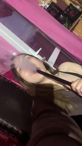 A young woman with long brown hair is in the process of smoking a hookah. She is holding the hookah hose in her right hand and bringing it to her mouth. Smoke is visible in the air around her face. She is wearing a dark top. The background appears to be indoors, with pink curtains and wooden walls. There are some speakers and decorations visible on the wall in the background. The lighting is somewhat dim.