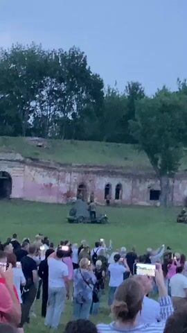 A wide shot captures an outdoor historical reenactment event as dusk settles. A large crowd of spectators, many holding up their phones to record, stand in the foreground. Behind them, a grassy field slopes upwards towards a long, weathered brick structure resembling a fort or barracks with arched openings and a moss-covered roof. Several period military vehicles, including what appears to be a tank or armored car, are positioned on the field. Figures dressed in historical military uniforms are visible around the vehicles and within the structure, engaged in the reenactment. Trees and foliage frame the upper portion of the scene, with a pale, overcast sky overhead. The overall impression is of a staged historical event being observed by a large audience.