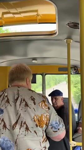 The video shows the interior of a yellow bus from a rear-facing perspective. In the foreground, a person with blonde hair is wearing a light-colored shirt with a floral pattern. They are seated, facing forward. To the left, another person is visible, wearing a plaid shirt. The bus has yellow poles for support and a yellow roof with a skylight. Through the windows, greenery and a road are visible, suggesting the bus is moving through a rural or wooded area. The lighting is natural and appears to be daytime. The overall scene conveys movement and travel.