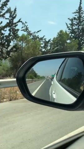 A white car is driving on a highway. The view is from inside the car, looking out the passenger side mirror. The mirror reflects the road ahead, with other cars visible in the distance. Green trees and foliage are visible on the side of the road, and the sky is blue and clear. The car appears to be moving at a moderate speed.