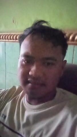 The video is a close-up selfie video of a man in a light-colored t-shirt. He has dark hair and a mustache. He is looking at the camera and smiling. The background appears to be a room with a green wall and some decorative molding. The lighting is somewhat dim. The video is short, only a few seconds long.