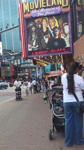 A street scene at daytime with a group of people walking on the sidewalk in front of various tourist attractions. A large billboard for "Movieland Wax Museum of the Stars" is visible, featuring images of movie characters. Several people are pushing strollers, and others are walking in groups. Cars are visible on the street in the background. The environment appears to be a busy tourist area with multiple businesses and colorful signage.