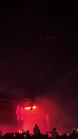 A recording taken at an indoor music concert shows a stage with red lighting. A dark figure or person stands or moves at the center of the stage structure, which features horizontal lighting fixtures. Two large, illuminated, white figures are visible on either side of the stage. The crowd is visible in silhouette near the bottom of the screen, with some people holding up phones. The overall atmosphere is dark and energetic, with the bright stage lights contrasting against the surrounding darkness.