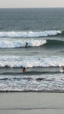 A drone-style shot shows a body of water with multiple waves rolling in towards the shore. The water is a deep blue-green color, and the waves are breaking with white foam. There are several people in the water. In the middle ground, one person is surfing on a wave, standing on a surfboard. In the foreground, closer to the shore, two people are in the water, one sitting on a surfboard and the other with their back to the camera, seemingly swimming or wading. Further out, beyond the surfer, two more surfers are visible, appearing to be paddling. The sky is a hazy light color, suggesting it's either early morning or late afternoon. The overall scene is a typical day at the beach with people enjoying the ocean.