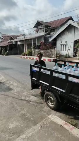 A man is seen unloading large plastic water jugs from the back of a small truck. He is wearing a black t-shirt and grey shorts, and flip-flops. He walks away from the truck carrying two jugs of water, one in each hand. The truck is parked on the side of a paved road, with a sidewalk in front of it. In the background, there are houses and buildings, as well as some power lines. The sky is cloudy.
