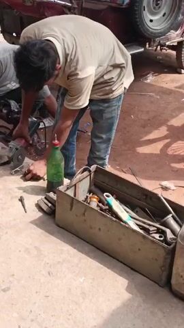 Two men are working on a vehicle outdoors. One man is using an angle grinder on a metal part, creating sparks. The other man appears to be working on a different part of the vehicle with tools. A toolbox and a green plastic bottle are on the ground nearby, along with other tools and components. The setting looks like a repair shop or garage.