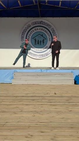 Two men stand on a stage in front of a circular banner that reads "FESTIVAL SEMEINOGO OTDYHA GARBATA PA-SVIAZKOUI MAGILEUSKI RAEN ROBAVOGO OBRAZA". The man on the left wears a teal suit and the man on the right wears a black suit. Both men have microphones and appear to be speaking or singing. The stage is made of wooden planks.