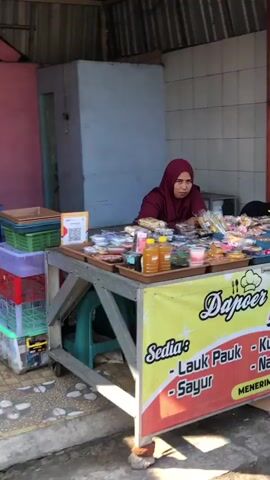 The video shows a woman wearing a hijab standing behind a stall filled with various food items. She is looking towards the camera. The stall is covered by a yellow banner that says "Dapoer" and lists various food items in Indonesian. Behind the stall, there is a grey structure, possibly a small room or shed, and a tiled wall. To the left of the stall, there are stacked crates. The ground appears to be paved. The lighting suggests it is daytime.