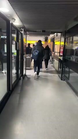 The video shows a hallway in an airport or similar building. The hallway is lined with glass doors and has a gray tiled floor and a ribbed ceiling. People are walking through the hallway in both directions. Some are pulling luggage. One man wears a bright yellow safety vest and walks directly towards the camera. The hallway opens into a larger waiting area. There are some kiosks and chairs visible in the waiting area. There are red signs on the doors with warnings in Spanish and English. The overall lighting in the hallway is bright.