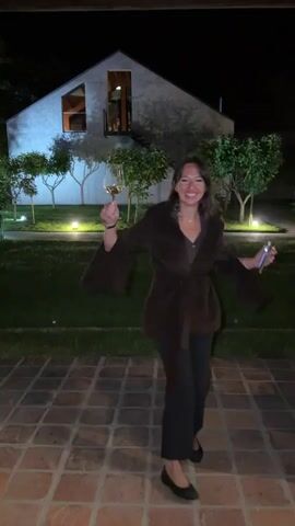 The video shows a woman at night standing on a brick patio in front of a modern house with a dark roof and white siding. She is wearing a brown velvet wrap, dark pants, and black flats. The woman holds a glass of white wine in one hand and a phone in the other. She appears to be celebrating, shouting and smiling, extending the wine glass towards the camera at one point. The background includes a lawn with several small trees illuminated by ground lights.