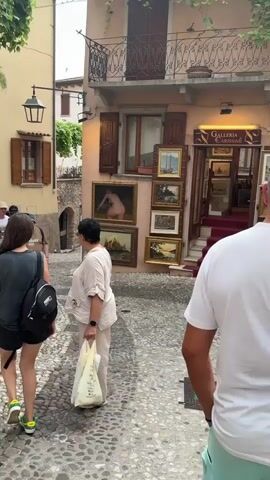 This video captures a bustling scene on a cobblestone street in Malcesine, Italy. Several people are walking, some appearing to be tourists. In the foreground, a person with a backpack is walking away from the camera, while another individual in a white shirt and shorts walks towards the viewer. To their left, a man in a brown t-shirt and blue shorts walks with his back to the camera, followed by another person.

The street is lined with buildings, one of which houses an art gallery named "GALLERIA CARISSIMI." Several paintings are displayed on the exterior wall of the gallery, including a nude figure, sailing boats, and landscapes. The gallery has a red carpet leading up to its entrance. The architecture suggests a historic European town.

The time of day appears to be daytime, with ample natural light, and the weather seems pleasant. The atmosphere is one of casual exploration, with people enjoying a walk through the charming Italian town. The scene is vibrant, with the colorful artwork and the activity of the pedestrians contributing to the lively ambiance.