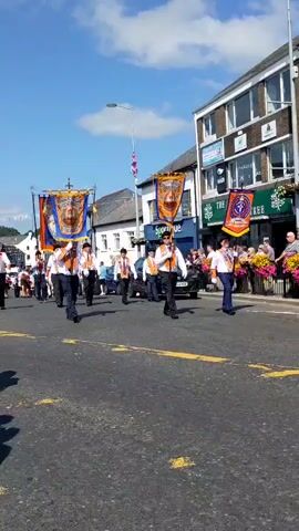 This is a first-person perspective video of a parade. The camera is positioned on a street with onlookers on the left side. In the foreground, there are yellow lines painted on the asphalt. The parade participants are walking in formation on the street, some of whom are carrying large banners. The participants are wearing white shirts, dark pants, and orange sashes. The banners are orange and blue and appear to have images and text on them. The buildings on the right side of the street have storefronts. The sky is blue and the weather appears to be sunny.