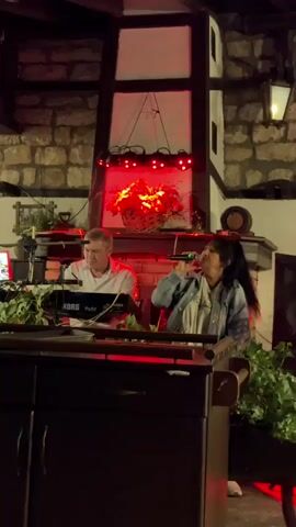 A man and a woman are performing music in front of a stone wall, likely a restaurant or event venue. The man sits at a keyboard, which appears to be a KORG, and is focused on playing. The woman stands to his right, holding a microphone and singing. They are positioned behind a dark wooden counter or podium, decorated with green foliage. Above them, a large, framed structure with a basket of red flowers sits on a mantelpiece. The lighting shifts from green to pink and back to green, illuminating the performers and the floral arrangement. The overall atmosphere suggests a live musical performance.