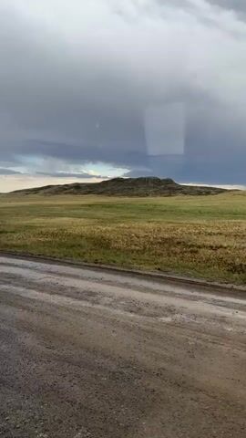 The video captures a landscape scene from a moving vehicle. The foreground shows a dirt road. Beyond the road is a wide expanse of grassy terrain, with dry, yellowish grass dominating the middle ground. In the background, a large, rocky hill or mesa rises, its surface appearing rough and uneven. The sky above is overcast with dark, dramatic clouds, suggesting impending rain or a stormy weather system. The overall impression is of a vast, open, and somewhat desolate natural environment.