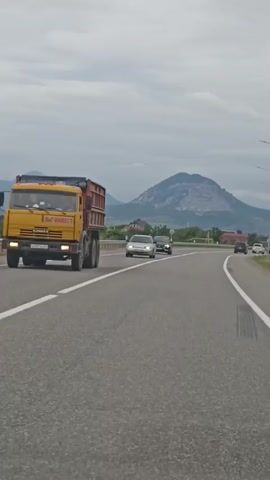 The video captures a car's perspective driving on a highway with mountains in the background. The highway has clear white lane markings, and guardrails are visible along the side. Several cars are on the road, including a silver sedan and a black car. In the distance, a distinctive, flat-topped mountain dominates the landscape, with other, more pointed mountains behind it. The scene appears to be in a rural or semi-rural area, with some houses and trees visible between the highway and the mountains. The sky is overcast, suggesting a cloudy or possibly rainy day. The overall mood is one of travel and exploration through a scenic, mountainous region. A sign with "Садовый" is visible, indicating the location is Sadovy, Russia.