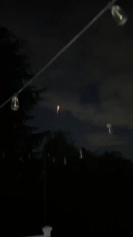 A short video of fireworks being set off at night. The fireworks are yellow and gold and explode in the dark sky. The video is shot from below, looking up at the fireworks. String lights are visible in the foreground, adding to the festive atmosphere. The sound of fireworks is heard throughout the video.