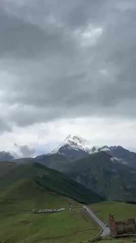 The video begins with a panoramic view of a vast mountainous landscape under a cloudy sky. The foreground shows rolling green hills. In the middle ground, a winding road is visible, with a cluster of buildings and parked cars. Further in the distance, a majestic snow-capped mountain peak dominates the horizon, partially obscured by clouds. The camera pans slightly, revealing more of the green valleys and rugged terrain. Towards the end of the video, the viewpoint shifts to a closer perspective of the road, showing several cars parked alongside it and a few people walking in the grassy area beside the road. The lighting suggests an overcast day, possibly late afternoon or early morning.
