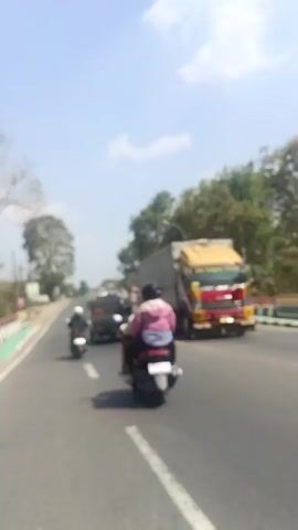 A blurred video shows a sunny day on a road. A motorcycle with a rider wearing a pink shirt is in the foreground, driving on the right side of the road. In the background, other vehicles, including a truck, are also visible. Trees line the sides of the road, and the sky is blue with some clouds. The video captures a moment of traffic on a clear day.