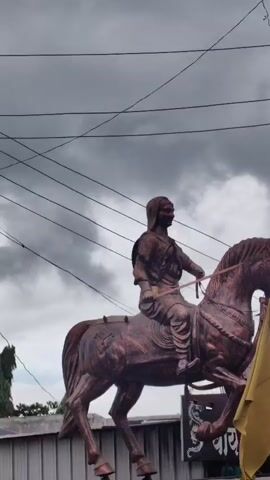 A bronze statue of a woman on horseback is shown from the waist up against a cloudy sky. The woman wears a head covering and traditional clothing, and she holds a sword in her right hand. The horse is also bronze and appears to be in mid-stride. A yellow flag is visible to the right of the statue. The statue is mounted on a pedestal that is not fully visible. The video is slightly shaky and zooms in on the statue.