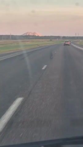 The video shows a road with cars and a power plant in the background. The sky is a pale pink and blue, suggesting it is either sunrise or sunset. The power plant has a tall smokestack emitting smoke. The landscape is rural with fields of grass and trees.