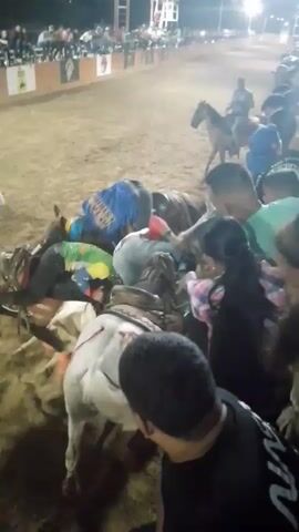 The video shows a horse-related competition, likely a rodeo, taking place in an arena with an audience in the background. Several people are gathered closely around multiple horses, some of which are wearing saddles and being mounted. People are wearing casual clothing, including t-shirts, jeans, and button-down shirts, with one rider sporting a Venezuelan flag-themed outfit. The ground appears to be dirt or sand, typical of a rodeo or horse-show arena, and the scene is illuminated by overhead lighting. One person is standing on a horse that has fallen to the ground. People are assisting that person, trying to help them off the horse. There is general activity and movement as the participants prepare for or are in the midst of the competition.