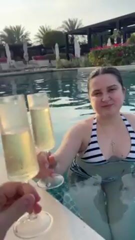 A person in a pool holds out a glass of champagne for a toast. The woman in the pool wearing a black and white striped swimsuit raises her own glass and takes a sip. She wears a key-shaped necklace. The pool is surrounded by lounge chairs, palm trees, bushes with pink flowers, and shaded pergolas. The pool tiles are blue and green. The time of day appears to be late afternoon or early evening.