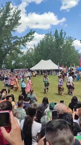 This video captures an outdoor cultural event, likely a powwow, with many Indigenous people dressed in traditional regalia. The foreground is filled with spectators holding up their phones to record the event. In the middle ground, several dancers are performing in a grassy field. They are adorned in colorful, elaborate outfits, some featuring feathers and intricate beadwork. A white tent is visible in the background, likely serving as a shaded area or vendor space. The scene is set under a bright blue sky with fluffy white clouds, suggesting a warm, sunny day. Trees form a backdrop to the gathering. The overall atmosphere appears festive and celebratory.
