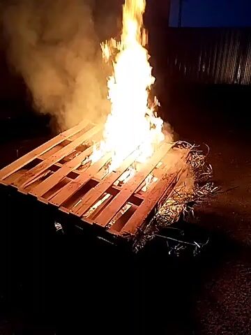 A wooden pallet is on fire in an outdoor space at dusk. The flames are tall and bright, accompanied by white smoke. The surrounding area is dark, with a building or fence visible in the background.