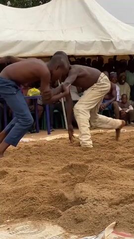 Two young boys wrestle in a sand pit while an audience watches. The boy on the left wears ripped beige pants and has no shirt, and the boy on the right wears a blue pair of pants and also has no shirt. They both grab onto one another and struggle for dominance. In the background, an audience watches under a white tent. A man in red pants walks behind the wrestlers.