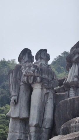 The video shows a group of stone statues. There are five figures standing close together on a stone base. The figures appear to be a mix of adults and children. One of the adults is wearing a hat, and two figures appear to be holding bundles. The background is hazy, but trees can be seen in the distance. The statues are gray and weathered.