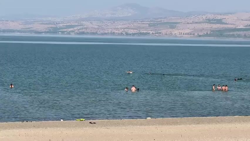 The video shows a wide shot of a body of water, likely a lake, with a sandy beach in the foreground. The water is calm and appears to be a deep blue color. In the middle ground, several people are swimming and wading in the water. On the beach, there are a few small objects, possibly litter or personal belongings. In the background, a hilly landscape stretches across the horizon under a hazy sky. The overall scene depicts a serene day at a waterfront.