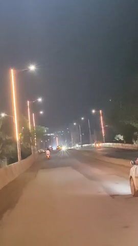 A night time video capture from a vehicle moving on a road. The road is lined with streetlights. The headlights of approaching vehicles are visible and cause lens flare. Trees are visible on the side of the road.