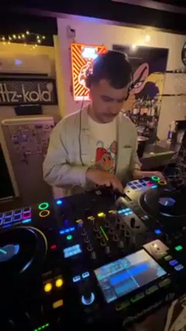 A DJ is working at his mixing board at a bar in a medium shot. He is a light-skinned male with a mustache wearing headphones, a white t-shirt, and a light green button up shirt. He is standing behind a black Pioneer DJ mixing board with multiple brightly lit buttons, knobs, and a screen.  He has his hands on the board, seeming to be in the process of mixing music, and looking at the screen and controls. In the background there is a neon sign on the wall and a shelf with various liquor bottles and glassware. The lighting in the bar is dim with purple and other colored lights illuminating the mixing board and the DJ. The camera is positioned at a medium angle, slightly below the DJ's eye level. At one point a hand appears briefly in front of the camera.