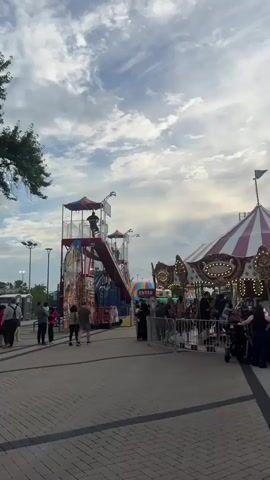 The video shows a lively carnival scene under a cloudy sky. In the foreground, a portion of a carousel with red and white striped awnings and illuminated decorations is visible, with people queuing to enter. To the left, a multi-story carnival ride with bright colors, including red, blue, and yellow, stands prominently. People are seen walking on a paved area in front of these attractions. The overall environment suggests a public gathering at an amusement park or fair.